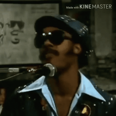 A Man Is Singing Into A Microphone Wearing Sunglasses And A Hat . GIF
