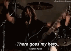 A Man Is Singing Into A Microphone While Holding A Guitar And A Drum Set . GIF
