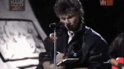 A Man Is Singing Into A Microphone While Holding A Notebook . GIF