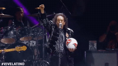 A Man Is Singing Into A Microphone While Holding A Soccer Ball On Stage . GIF