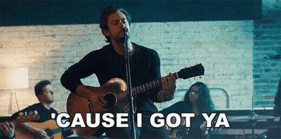 A Man Is Singing Into A Microphone While Playing A Guitar In Front Of A Brick Wall . GIF