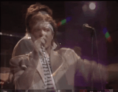 A Man Is Singing Into A Microphone While Playing A Guitar On Stage GIF