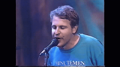 A Man Is Singing Into A Microphone While Wearing A Blue Shirt That Says Minutemen . GIF