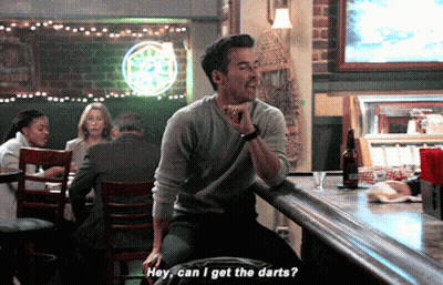 A Man Is Sitting At A Bar Talking To Another Man Who Is Asking If He Can Get The Darts GIF