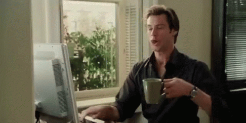 A Man Is Sitting At A Desk In Front Of A Computer Drinking Coffee . GIF