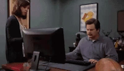 A Man Is Sitting At A Desk Using A Computer While A Woman Looks On . GIF