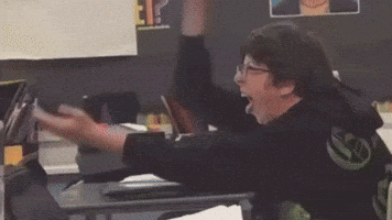 A Man Is Sitting At A Desk With His Arms In The Air Wearing A Black Shirt With The Letter W On It GIF