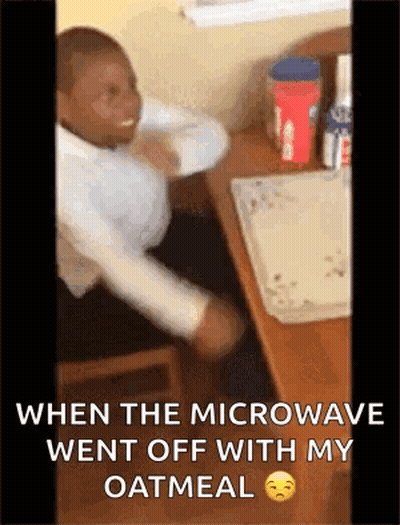 A Man Is Sitting At A Desk With His Arms Outstretched While A Microwave Went Off With His Oatmeal GIF