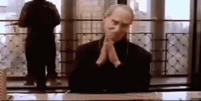 A Man Is Sitting At A Desk With His Hands Folded In Front Of A Window . GIF