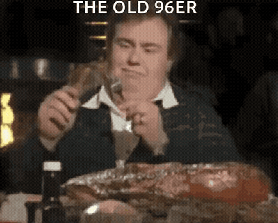 A Man Is Sitting At A Table Eating A Large Piece Of Meat While Holding A Fork . GIF