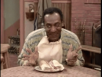 A Man Is Sitting At A Table Eating A Sandwich With A Bag Around His Neck . GIF
