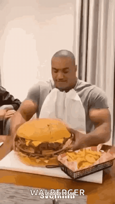 A Man Is Sitting At A Table Eating A Very Large Hamburger And French Fries . GIF