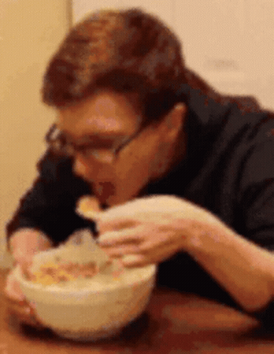 A Man Is Sitting At A Table Eating Cereal From A Bowl . GIF