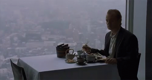 A Man Is Sitting At A Table Eating Food With Chopsticks And Looking Out A Window . GIF