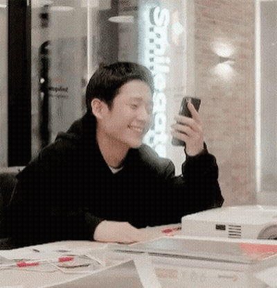 A Man Is Sitting At A Table Holding A Cell Phone And Smiling GIF