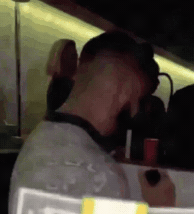 A Man Is Sitting At A Table In A Dark Room With A Red Cup In His Hand . GIF