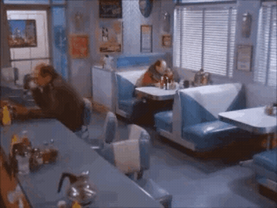 A Man Is Sitting At A Table In A Diner With Blue Booths . GIF