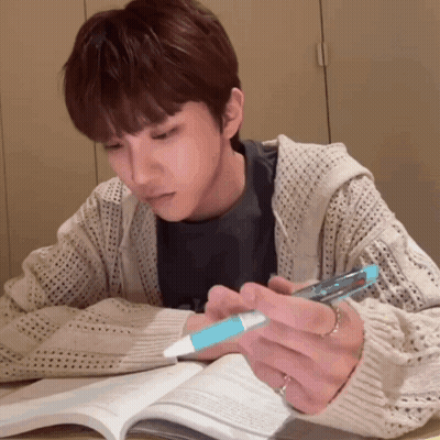 A Man Is Sitting At A Table Reading A Book And Holding A Pen GIF