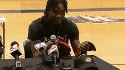 A Man Is Sitting At A Table Surrounded By Microphones Holding A Hat . GIF