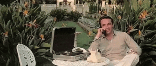 A Man Is Sitting At A Table Talking On A Telephone In A Garden . GIF