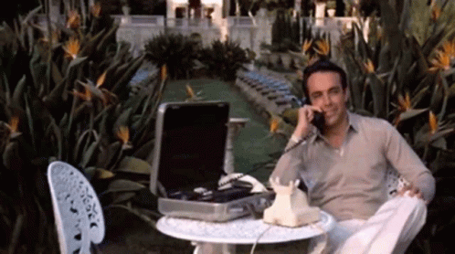 A Man Is Sitting At A Table With A Briefcase And Talking On A Phone GIF