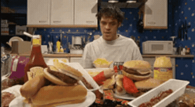 A Man Is Sitting At A Table With A Bunch Of Food Including Hamburgers And Hot Dogs .. GIF