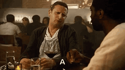 A Man Is Sitting At A Table With A Glass Of Wine And Talking To Another Man In A Restaurant . GIF