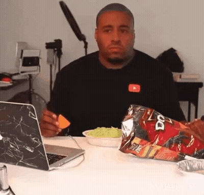 A Man Is Sitting At A Table With A Laptop And A Bag Of Doritos . GIF