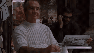 A Man Is Sitting At A Table With A Newspaper That Says Commentatore On It GIF