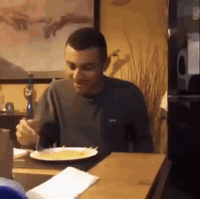 A Man Is Sitting At A Table With A Plate Of Food And A Fork In His Hand . GIF