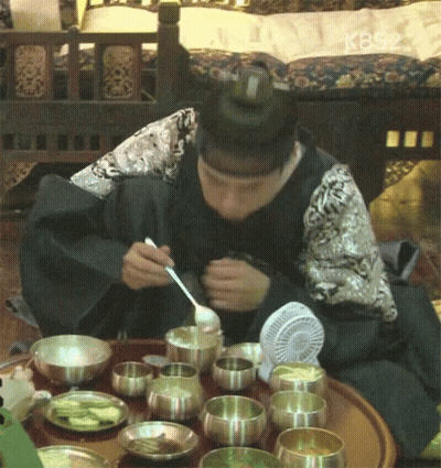 A Man Is Sitting At A Table With Bowls Of Food And A Fan And Eating With A Spoon And Fork GIF