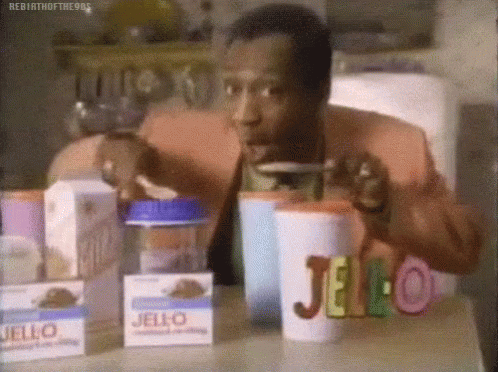 A Man Is Sitting At A Table With Boxes Of Jello And Drinking From A Cup . GIF