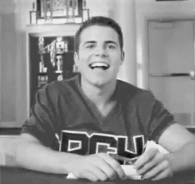 A Man Is Sitting At A Table With His Mouth Open And Smiling . He Is Wearing A Shirt With The Word Dru On It . GIF