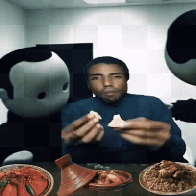 A Man Is Sitting At A Table With Plates Of Food And Two Mascots Behind Him GIF