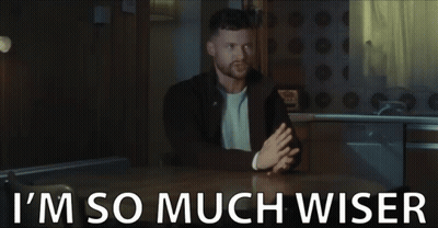 A Man Is Sitting At A Table With The Words `` I 'M So Much Wiser '' Written On The Bottom . GIF