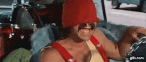 A Man Is Sitting In A Car Wearing A Red Hat And Overalls . GIF