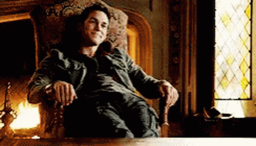 A Man Is Sitting In A Chair In Front Of A Fireplace And A Stained Glass Window . GIF