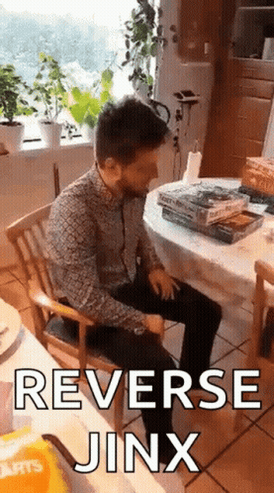 A Man Is Sitting In A Chair In Front Of A Table With A Stack Of Books And The Words Reverse Jinx On It . GIF