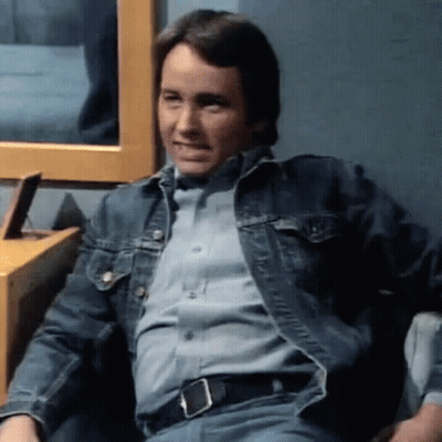 A Man Is Sitting In A Chair Wearing A Denim Jacket And A Blue Shirt . GIF