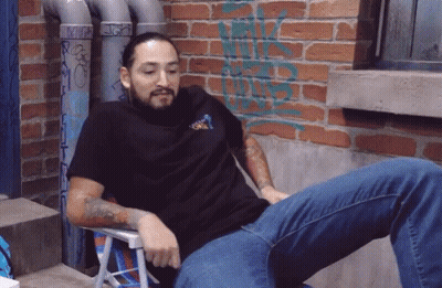 A Man Is Sitting In A Chair With A Brick Wall Behind Him With Graffiti On It That Says ' Rick ' GIF