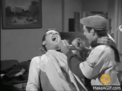 A Man Is Sitting In A Chair With His Mouth Open While Another Man Shaves His Beard . GIF