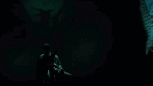A Man Is Sitting In A Dark Room With A Statue Of A Demon In The Background . GIF