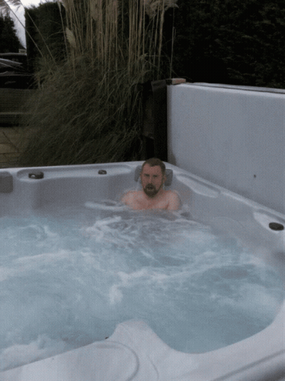 A Man Is Sitting In A Hot Tub With A Plant In The Background GIF