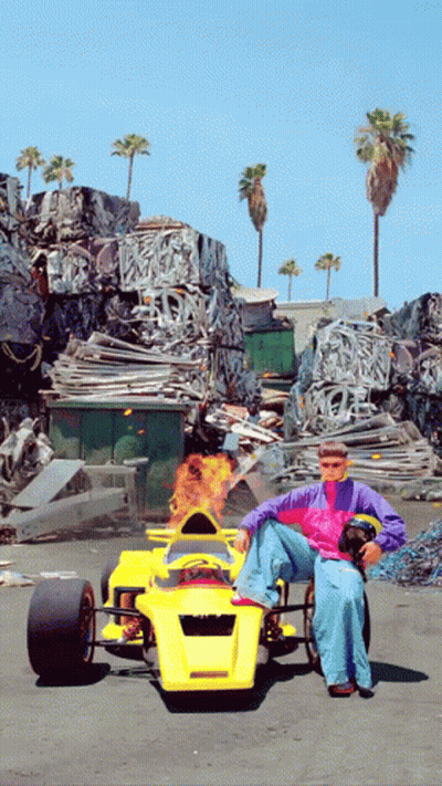 A Man Is Sitting In A Yellow Race Car With Flames Coming Out Of The Steering Wheel GIF