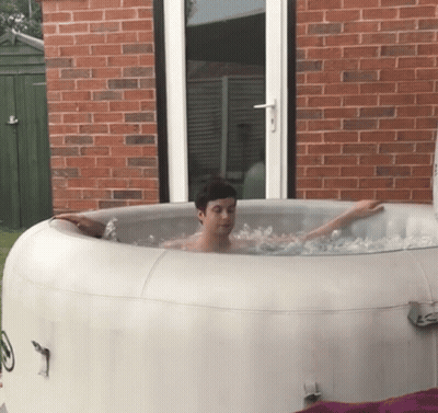 A Man Is Sitting In An Inflatable Hot Tub Outside Of A Brick House GIF