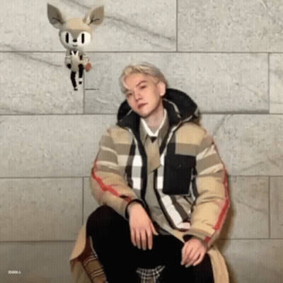 A Man Is Sitting In Front Of A Brick Wall With A Stuffed Animal Hanging From His Head . GIF