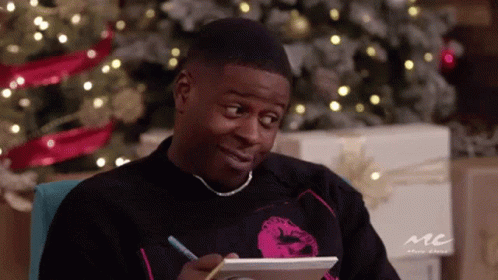 A Man Is Sitting In Front Of A Christmas Tree Writing On A Piece Of Paper With A Pencil . GIF
