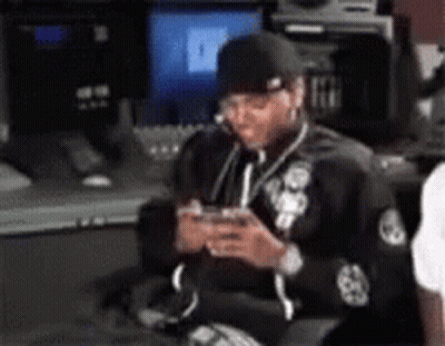 A Man Is Sitting In Front Of A Computer And Looking At His Phone . GIF