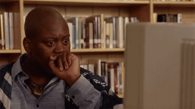 A Man Is Sitting In Front Of A Computer In A Library With His Hand On His Chin . GIF