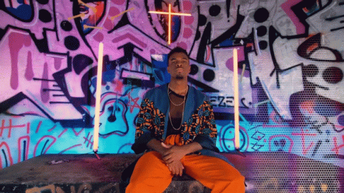 A Man Is Sitting In Front Of A Wall With Graffiti On It That Says Ezz GIF